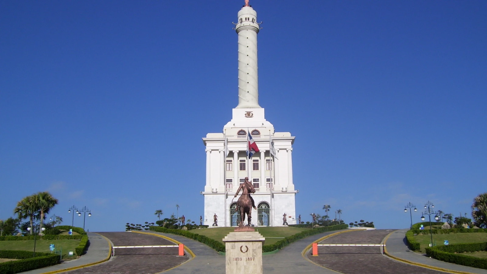 El Monumento a los Héroes de la Restauración en Santiago, con su torre blanca, la estatua en la cúspide y una estatua de bronce al frente
